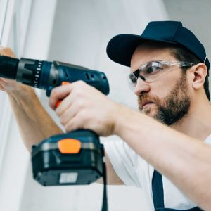 adult repairman in goggles fixing window handle by electric drill