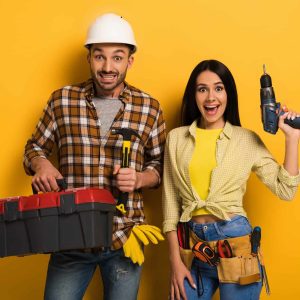excited handywoman and handyman holding toolbox and electric drill on yellow